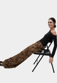 Svart off-shoulder topp och bruna byxor med leopardtryck i vida ben. Modell sitter på en svart hopfällbar stol, med mjukt tyg och en avslappnad passform.