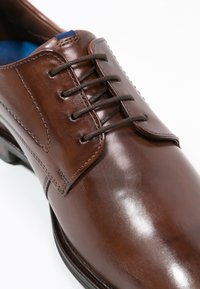 Chaussure en cuir marron avec une finition lisse, six œillets avec des lacets marron, et une languette texturée. Doublure intérieure bleue visible.