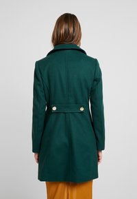 Cappotto di lana verde scuro con una grande colletto, silhouette aderente e accenti a doppio bottone sul retro, con una texture liscia.