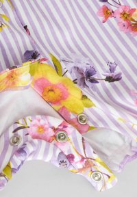 Combinaison florale en violet clair avec des rayures blanches, ornée de motifs floraux colorés, en matériau en coton, et dotée de boutons à pression métalliques au bas.