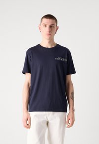 Marineblaues Baumwoll-T-Shirt mit Rundhalsausschnitt und kurzen Ärmeln, mit einem weißen Logo-Druck auf der linken Brust. Kombiniert mit weißen Hosen.