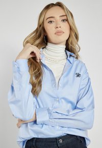 Jasnoniebieska koszula z długim rękawem, zapinana na guziki, z logo na piersi, uszyta z gładkiej tkaniny. Stylizowana na białym golfie.