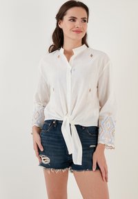 Blouse blanche à manches longues avec un nœud à l'avant, ornée de broderies géométriques dorées et bleues sur les manches et d'accents de diamants décoratifs.