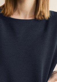 Kvinna med rakt axellångt hår som bär en mörk marinblå ribbad texturertopp med rund i halsringning.
