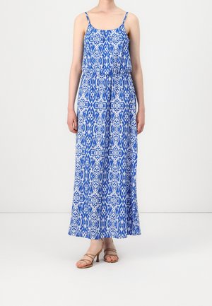 Femme portant une robe maxi sans manches à motifs bleu et blanc avec de fines bretelles et des sandales à talons hauts beiges à brides.