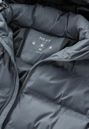 Interno di una giacca nera trapuntata con un'etichetta che presenta il marchio "NEXT", taglia "M" e icone per le caratteristiche trapuntate, impermeabili e di calore.