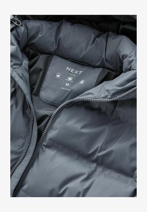 Musternikku musta jakk, mille sees on silt, millel on bränd "NEXT", suurus "M" ja ikoonid, mis näitavad jakkide, vihmakindlate ja soojustusomaduste kohta.