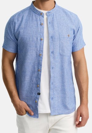 Hombre con camisa de manga corta azul claro con cuello mao, sobre camiseta blanca y pantalones blancos, mano en el bolsillo.