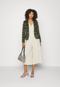 Blazer de patrón de cebra verde y negro sobre un mono beige claro, combinado con un bolso de rayas de cebra y mules de tacón abierto en lavanda.