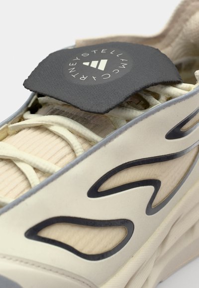 Zapatilla beige con superposiciones negras en forma de ola. Presenta un parche de logo redondo y texturizado y cordones claros sobre un material suave y ondulado.