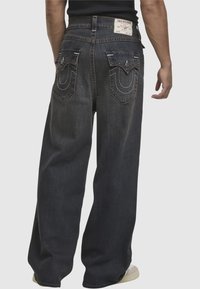 Ciemnoniebieskie dżinsy mają luźny krój, szerokie nogawki i charakterystyczne białe przeszycia na tylnych kieszeniach z metką marki na talii.