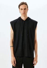 Sudadera sin mangas negra con cordón, textura suave y corte relajado. Presenta un tatuaje en el brazo y un fondo liso.