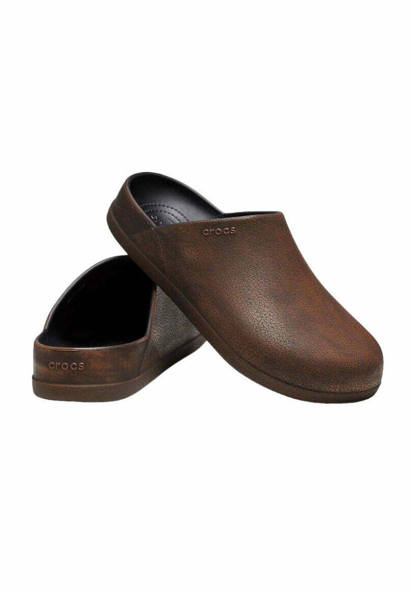 Bruine klompen van gestructureerd materiaal, met een instapontwerp en een ronde neus. Geëmbosseerd logo aan de zijkant. Zwarte binnenvoering.