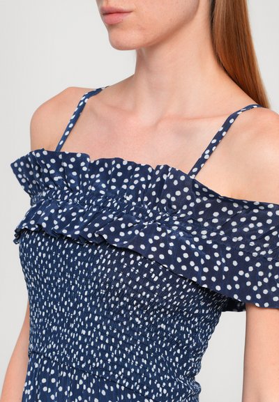 Marineblauwe off-shoulder jurk met witte stippen, met een ruches lijfje en geraffineerde textuur. Dunne schouderbandjes maken het ontwerp compleet.