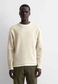 Beige strukturierter Sweatshirt mit Rundhalsausschnitt, langen Ärmeln, über einem weißen Hemd getragen, kombiniert mit olivgrünen Hosen.