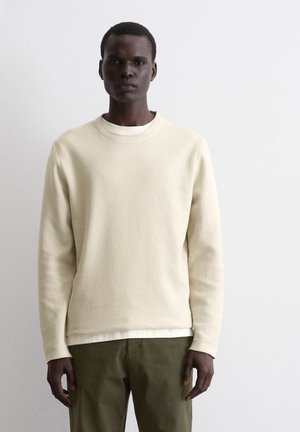 Beige textured sweatshirt with a round neck, long sleeves, layered over a white shirt, paired with olive green pants.