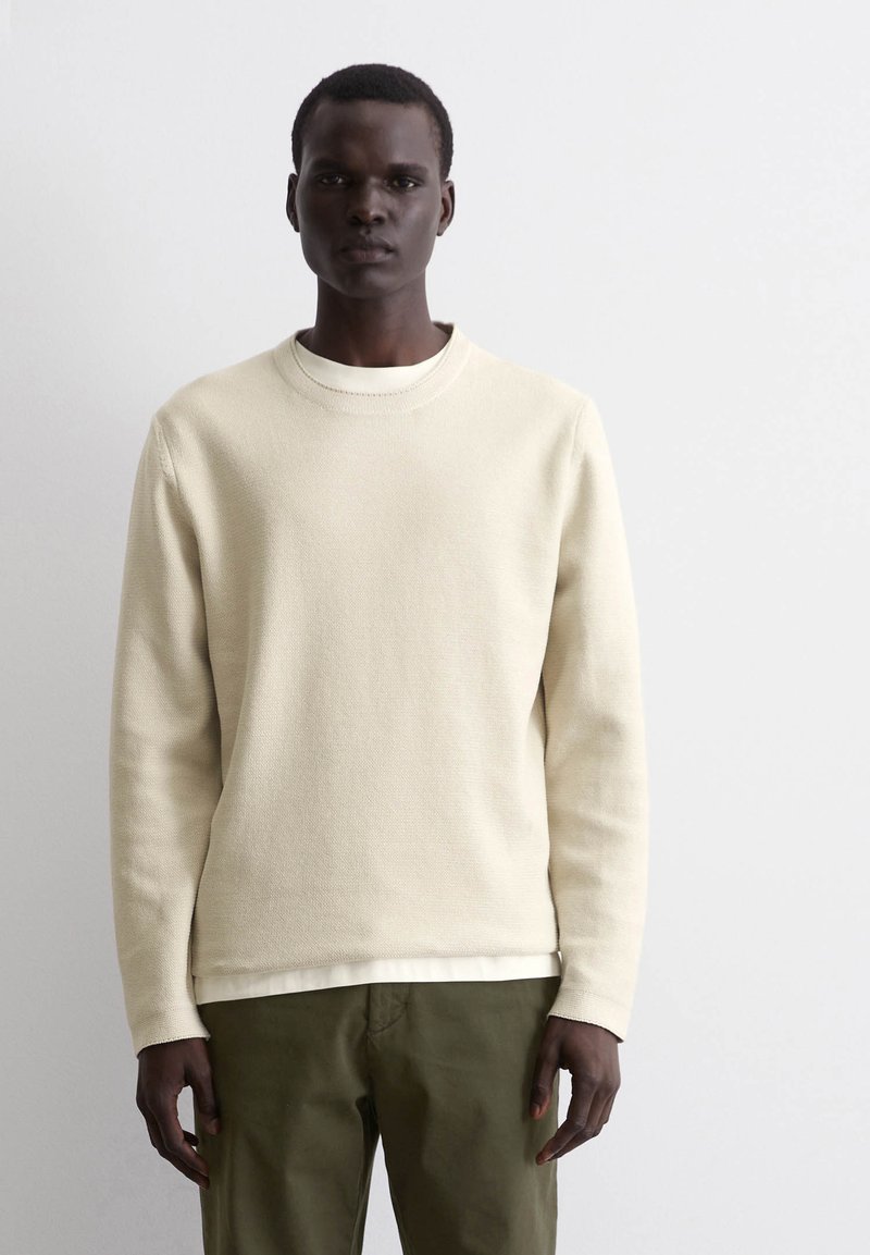 Beige strukturierter Sweatshirt mit Rundhalsausschnitt, langen Ärmeln, über einem weißen Hemd getragen, kombiniert mit olivgrünen Hosen.