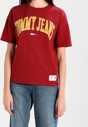 Red cotton t-shirt featuring "TOMMY JEANS" in bold yellow letters, with a small logo on the chest and a woven label at the hem.
