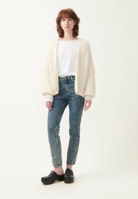Cream knit cardigan, loose fit; white long-sleeve shirt; faded blue high-waisted jeans; black wooden clogs. Simple, textured, and casual look.