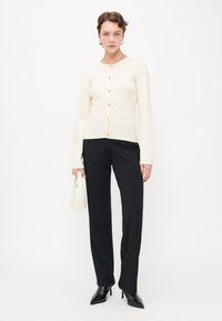 Ivory sweter z dzianiny z okrągłym dekoltem, sześcioma guzikami z przodu, dwoma kieszeniami z przodu, zestawiony z czarnymi spodniami i czarnymi szpilkami.