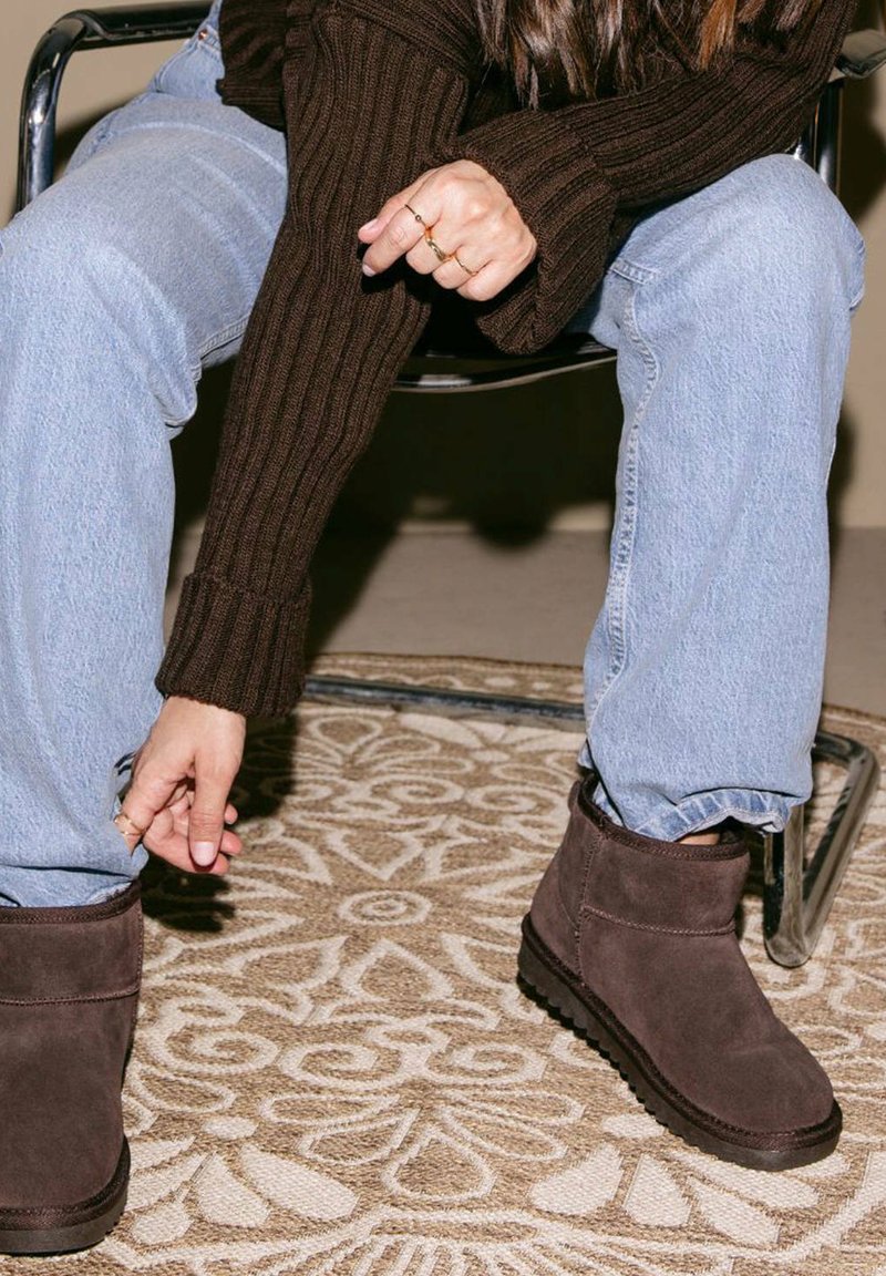 Bruine suède enkelboots met een gestructureerde afwerking, gedragen over lichtblauwe jeans terwijl je op een gemêleerd beige tapijt zit.