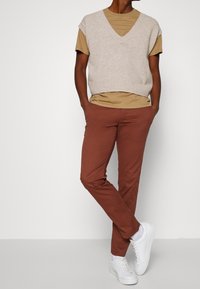 Beige knitted vest with a V-neck, layered over a tan T-shirt. Brown fitted pants and white sneakers complete the outfit. Smooth textures and simple design.