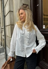 White cotton blouse featuring a ruffled front and lace trim on cuffs, paired with dark jeans and a brown leather bag.