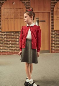 Red cardigan with frill accents over a white collared shirt, paired with a grey skirt. Black shoes with white ankle socks. Background features wooden panels.