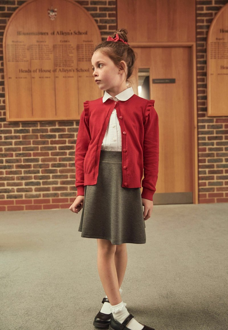 Red cardigan with frill accents over a white collared shirt, paired with a grey skirt. Black shoes with white ankle socks. Background features wooden panels.