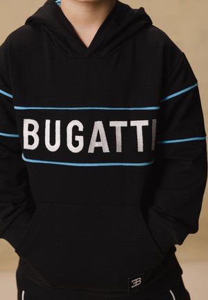 Niño con sudadera negra con rayas azules y grande texto blanco "BUGATTI" en el pecho, manos en el bolsillo delantero, fondo beige.