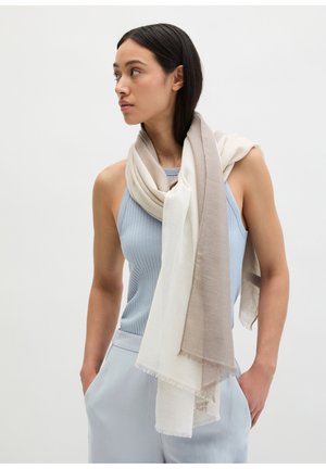 Junge Frau in hellblauem ärmellosem Top und Hose trägt einen zweifarbigen beigen und weißen Schal locker über die Schultern gelegt und schaut nach links.