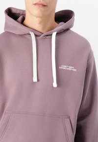 Bluza z kapturem z miękkiego, lawendowego materiału. Posiada białe sznurki, kieszeń z przodu i nadrukowany tekst: "CORE DEPT. ZAŁOŻONA 1990."