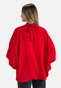 Rote, strukturierte Bluse mit einer Schleife am Hals und weiten Ärmeln. Der Rücken zeigt einen tiefen Ausschnitt und ein drapiertes Stoffdesign.