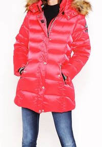 Parka matelassée rouge avec une capuche doublée de fourrure, des fermetures à pression et des poches latérales. Présente des accents rayés noir et blanc sur les manches et le col.