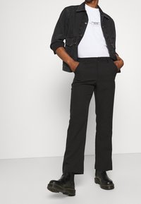 Osoba ubrana w czarną krótko skrojoną kurtkę na białej koszulce z nadrukiem, czarne spodnie o prostych nogawkach oraz czarne masywne botki, z rękami w kieszeniach.