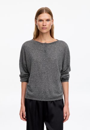 Femme aux cheveux blonds et raides portant un pull gris à manches longues boutonné et un pantalon noir, debout devant un fond blanc.