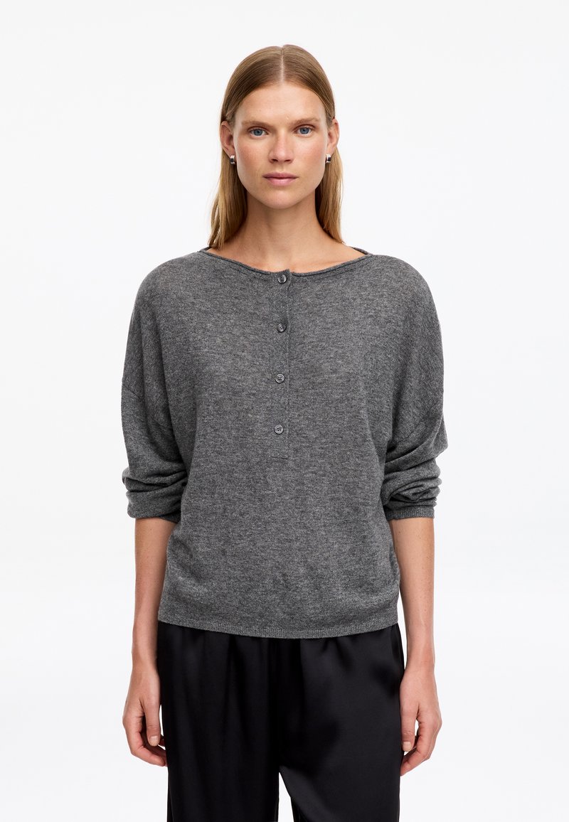 Femme aux cheveux blonds et raides portant un pull gris à manches longues boutonné et un pantalon noir, debout devant un fond blanc.