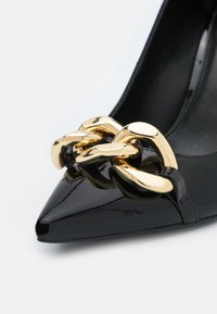 Chaussure à talon haut en cuir verni noir avec un bout pointu et un accent en chaîne dorée proéminente à l'avant. Texture lisse et design épuré.