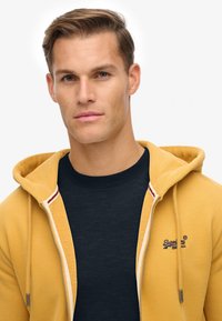 Giovane uomo con capelli corti castani, indossa una camicia blu navy e una felpa gialla senape con zip e un logo sul petto, che guarda di fronte.