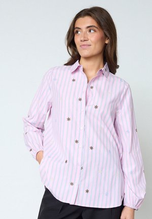 Kvinde iført en lyserød og blå striped knap-op skjorte med små broderede blomster, hænderne i lommerne, kigger lidt til siden.