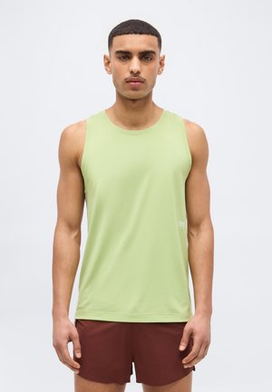 Νέος άντρας με κοντά μαλλιά φοράει ανοιχτό πράσινο αμάνικο αθλητικό μπλουζάκι και μπορντό σορτς για τρέξιμο, στέκεται μπροστά σε απλό φωτεινό φόντο.