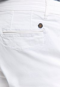 Witte broek met een achterzak, voorzien van een klein hexagonaal zwart en gouden accent. Glad materiaal met een eenvoudig, strak design.