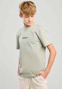 Camiseta de algodón de color verde claro con mangas cortas, que presenta un texto gráfico negro "INTO THE WILD" en el frente. Ajuste casual, textura suave.