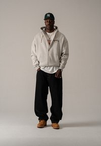 Sivá mikina na zips s logom, nosená cez biele tričko, doplnená voľnými čiernymi džínsami a hnedou obuvou. Zelená baseballová čiapka.