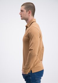 Brązowa koszulka polo z długim rękawem wykonana z miękkiego materiału, o luźnym kroju, z bocznymi rozcięciami i klasycznym kołnierzykiem, zestawiona z niebieskimi dżinsami.