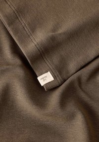 Brązowa tkanina o gładkiej fakturze, z zagiętym brzegiem i małą metką. Materiał wydaje się rozciągliwy i lekki.