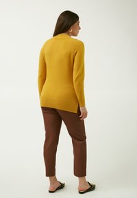 Maglione giallo senape con collo alto, orlo a costine e maniche lunghe, abbinato a pantaloni marroni e sandali neri piatti con borchie.
