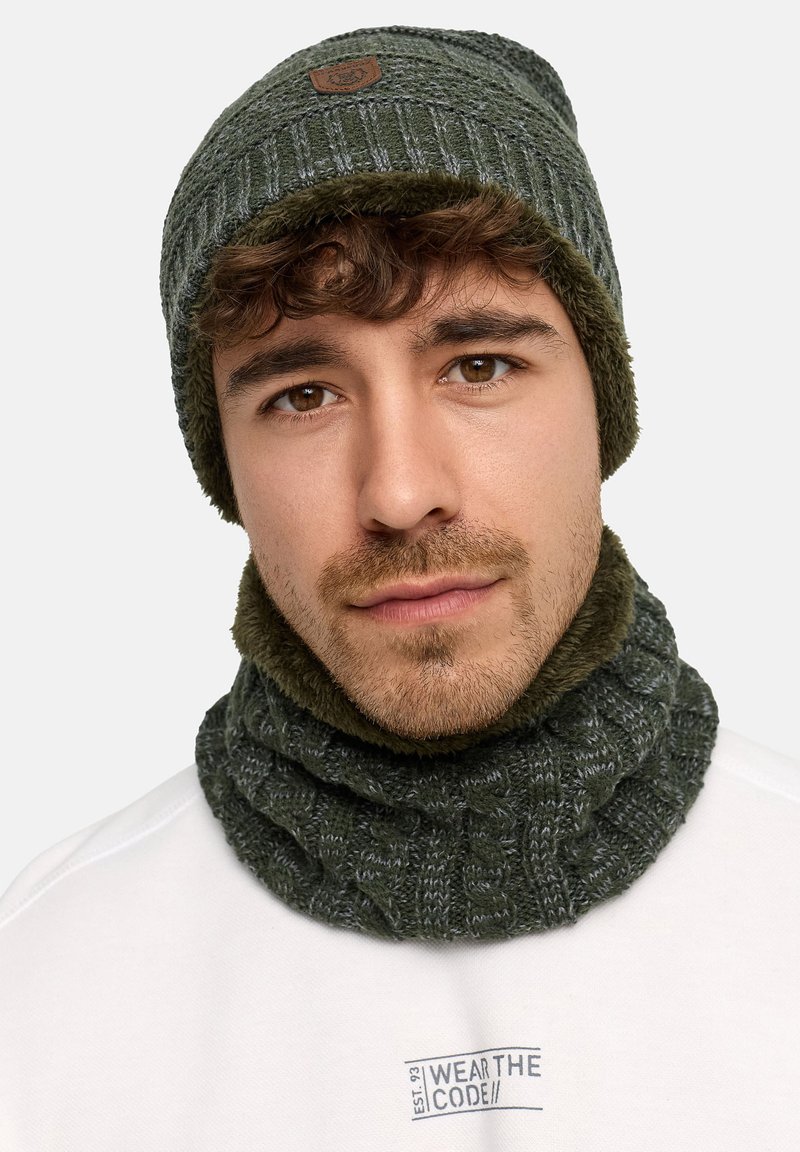 Cappello di lana verde scuro con motivo a coste e targhetta di pelle marrone, abbinato a una sciarpa a trecce dello stesso colore con una texture morbida.