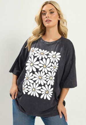 Frau mit langen blonden Haaren trägt ein übergroßes schwarzes T-Shirt mit weißem Gänseblümchenmuster und blaue Jeans vor einfarbigem Hintergrund.
