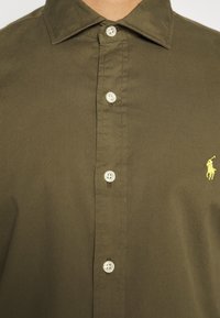 Camicia verde oliva scuro con bottonatura in cotone, dotata di colletto classico e due chiusure a bottone. Presenta un piccolo logo giallo sul petto.
